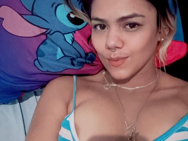 TarahDustin with pouty lips, nose ring, and blonde hair, wearing a striped top, showing her tits, and a necklace, lying on a blue and purple pillow.