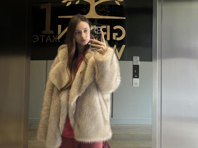 LaraSweetyHot met lang bruin haar, gekleed in een pluizige beige jas over een rode jurk, die een spiegelzelfportret maakt met een telefoon, waarbij haar fronsende gezicht en subtiele make-up te zien zijn..