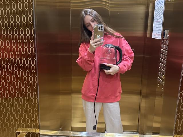 LaraSweetyHot with long brown hair, wearing a pink hoodie and white pants, holding a phone and a black corded appliance in a gold elevator.