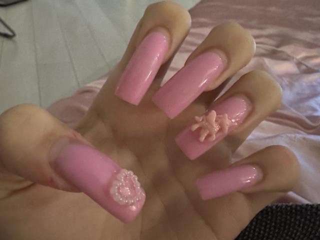La main de LaraSweetyHot, avec de longs ongles roses brillants, dont un est décoré d'une fleur et d'un cœur roses. Ses fesses ne sont pas visibles sur l'image..