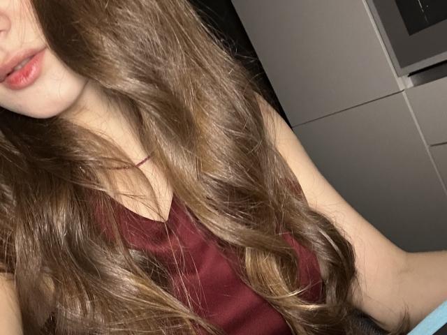 LaraSweetyHot with long, wavy brown hair, red tank top, and pink lips, showing her chest and part of her arm. Boobs visible, but not the focus.