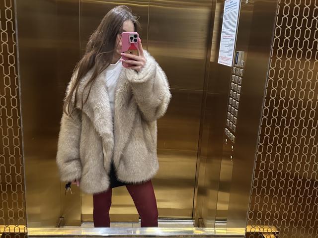 LaraSweetyHot draagt een pluizige beige bontjas, een witte T-shirt en rode broek, en maakt een spiegelzelfportret in een gouden lift, terwijl ze een roze iPhone met een rode hoes vasthoudt..