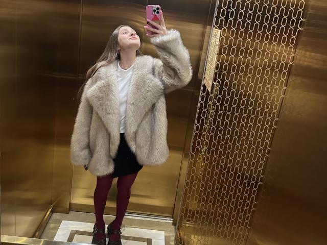 LaraSweetyHot with fluffy beige fur jacket, white shirt, black mini-skirt, red tights, and red heels, taking a selfie in a gold elevator, holding a pink phone.