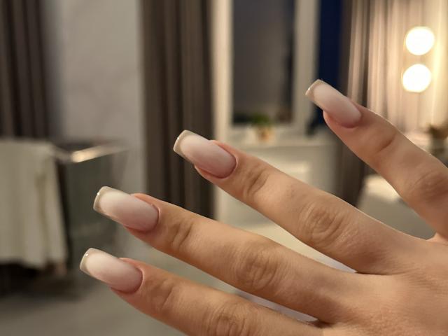 De hand van LaraSweetyHot, met lange, bleekroze nagels met een Franse tip. Haar vingers zijn licht gespreid, waardoor haar netjes gemanicuurde nagels in een zacht, warm licht worden getoond..