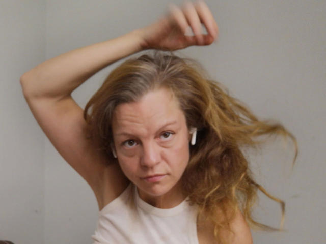 HotLena1984-sex has messy blonde hair, raised arm, white earbuds, and a serious face wearing a sleeveless white top. Her tits are small, and she's looking at the camera.