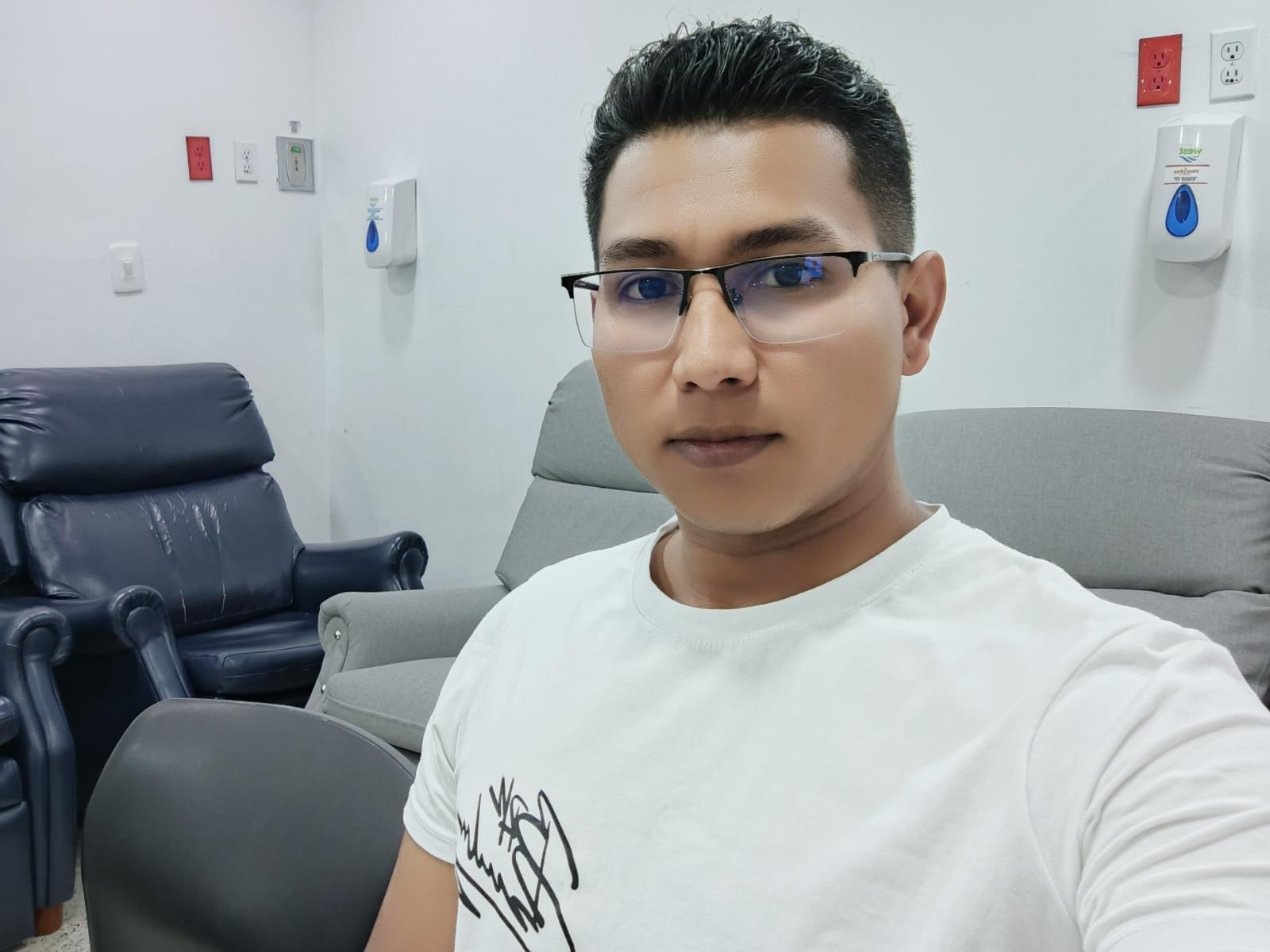 NicolaysRico has short black hair, blue glasses, and a medium build with light brown skin, wearing a white shirt. He's sitting in a gray office chair.