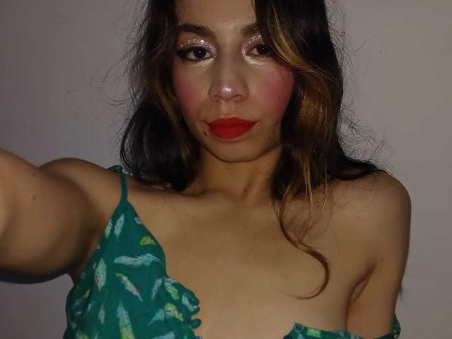 KatherinJohns with shiny eyes, red lipstick, and a green floral top, exposing her small tits, with wavy brown hair, extending her arm for a selfie.