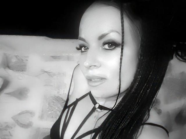 Black-and-white selfie of EmillySun-hot with long braids, dark eyeliner, and strappy top, showing her tits and a subtle smile, wearing a necklace. Her eyes are half-open.