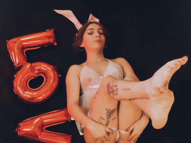 AndyDupont in white lace lingerie with bunny ears, legs up, showing tattoos, and red balloon "5" in the background. She's got a cute face and a sexy look.