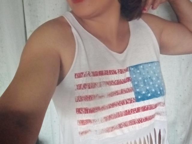 A camiseta branca de Rahel69, com um desenho da bandeira americana em vermelho e azul, mostra seus ombros e braço descobertos, com o rosto parcialmente fora do enquadramento..