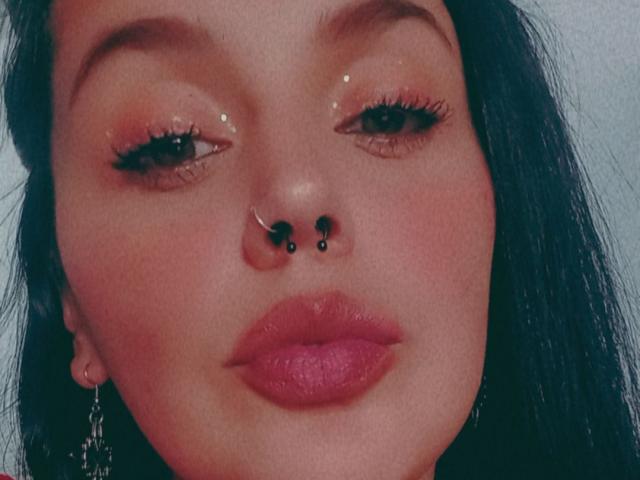 IsyMoon has shiny eyes with black eyeliner, a silver nose ring with black studs, and pink glossy lips. She has dark hair and a pink earring.