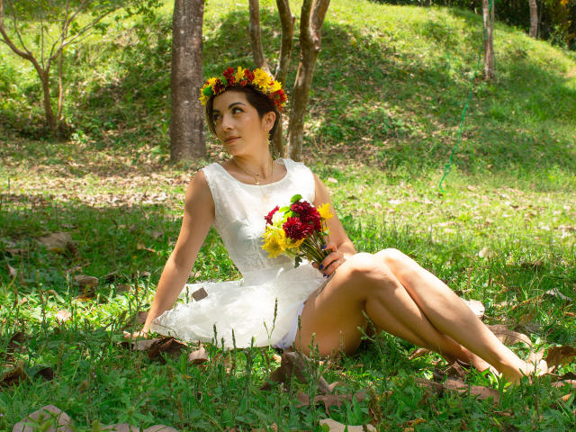 ChanelJolie is a sexy woman with brown hair, wearing a white dress and flower crown, sitting on grass, holding a colorful bouquet, showing her tits and ass.