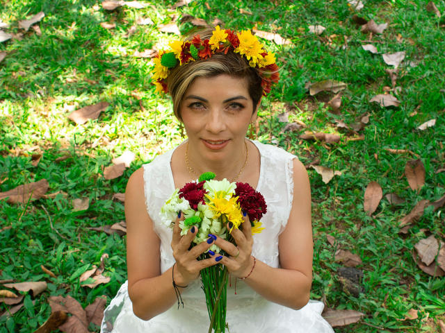 ChanelJolie with short blonde hair, wearing a flower crown and white dress, holding a bouquet of yellow, red, and white flowers, sitting on green grass with leaves.