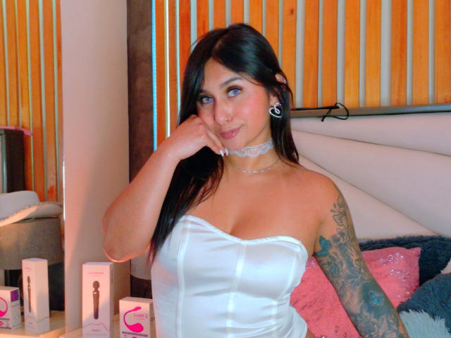 SarahOriz has dark hair, blue eyes, white strapless top showing her tits, choker, tattooed arm, and black ear gauges, sitting with hand on cheek, smiling.