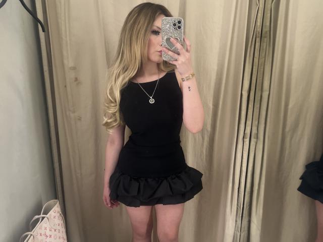 BellBlonde with blonde hair, black top, and black skirt, holding phone, showing off her ass, wearing a necklace and bracelet, standing in front of a mirror.