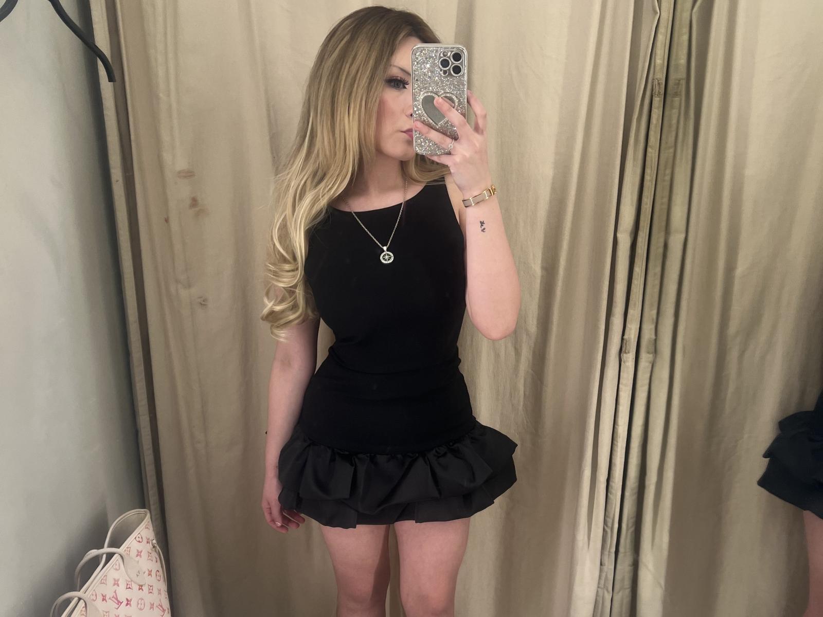 BellBlonde with blonde hair, black top, and black skirt, holding phone, showing off her ass, wearing a necklace and bracelet, standing in front of a mirror.
