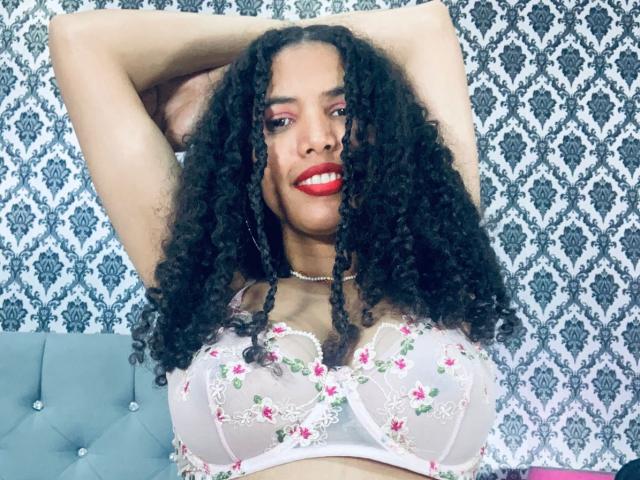 ZoeJonees with curly black hair, red lipstick, floral bra, arms raised, smiling, showing large boobs, wearing a pearl necklace, against a patterned background.