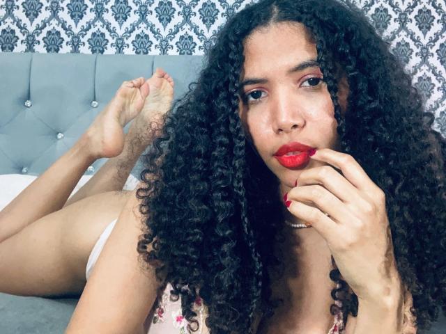 ZoeJonees with curly black hair, red lipstick, and red nails, lying on a bed, legs up, wearing white underwear, looking at camera, hand near mouth, soles visible.