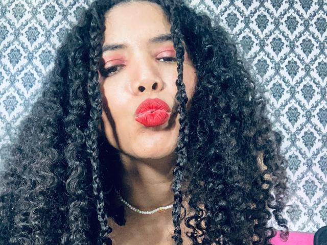 ZoeJonees with curly black hair, red lipstick, and a pearl necklace, pouting her full lips. She's wearing pink eyeshadow and has a medium brown skin tone.