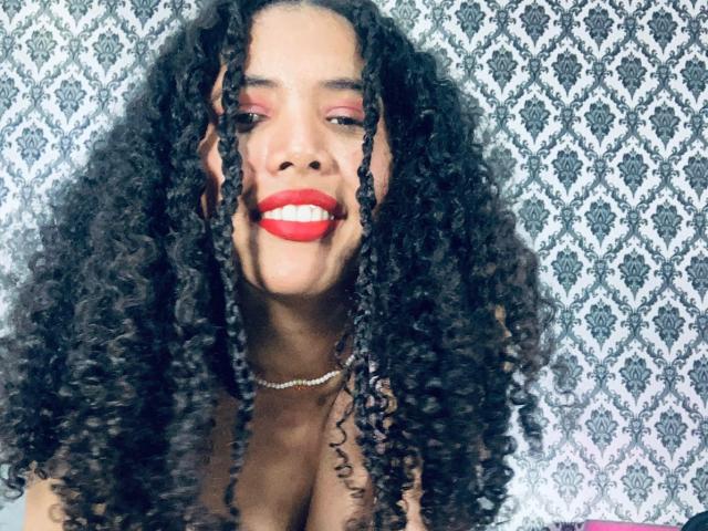 ZoeJonees with curly black hair, red lipstick, and a beaded necklace, smiling with pink eyeshadow. Boobs not visible, just her face and upper chest.