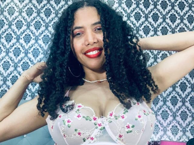 ZoeJonees with curly black hair, red lipstick, and white floral lace lingerie, showing her big tits and smiling. She's wearing a pearl necklace and large hoop earrings.