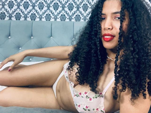 ZoeJonees with curly black hair, red lipstick, wearing a floral bra and white panties, lying on a bed with a tufted gray headboard, displaying her tits and ass.