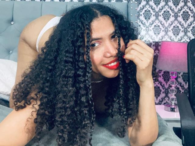 ZoeJonees with curly black hair, red lipstick, white panties, and a cute smile, on her knees, looking at the camera, her ass up, in a bedroom setting.