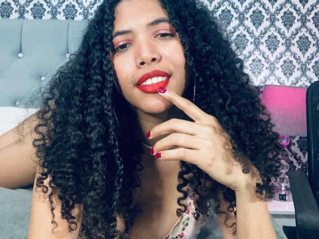 ZoeJonees with curly black hair, red lipstick, and red nail polish, gently touching her lips with her index finger, smiling, wearing a floral top, in a bedroom.