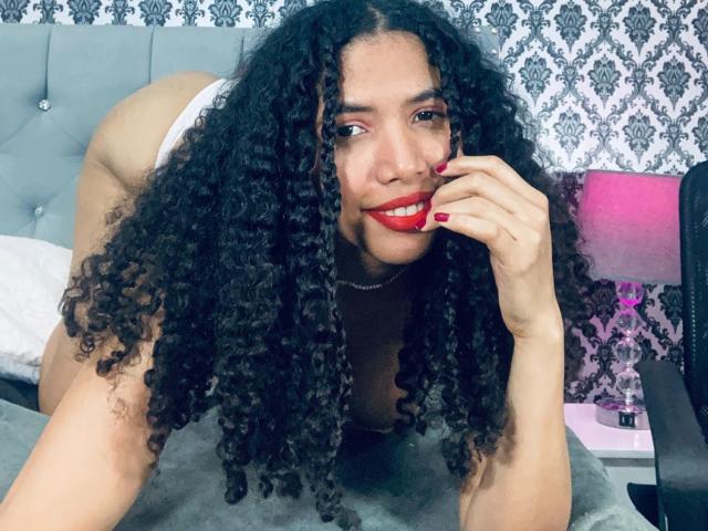 ZoeJonees with curly black hair, red lipstick, and white panties, on all fours, smiling, red nail polish, hand near mouth, on a gray bed, in a bedroom.