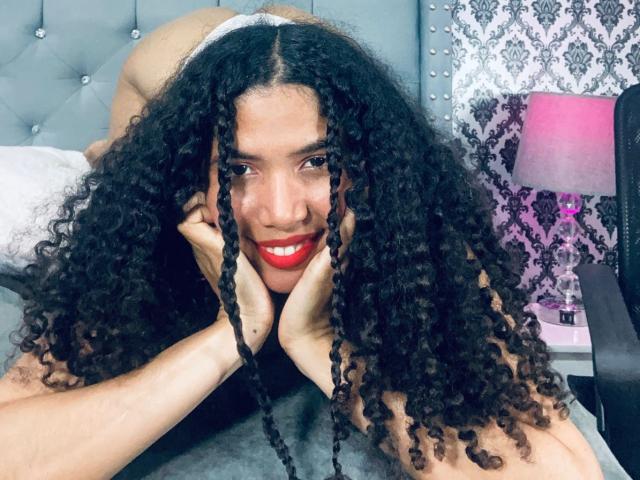 ZoeJonees with long, curly black braids, red lipstick, and smiling, hands on cheeks, lying on a bed, wearing white panties, her ass slightly raised. Bedroom setting, pink lamp.