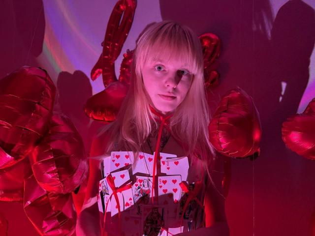Lialit, con cabello rubio, globos rojos y una camiseta con motivos de corazones, mirando directamente a la cámara, rodeada de puños hechos con globos rojos, bajo una iluminación rosa-roja..