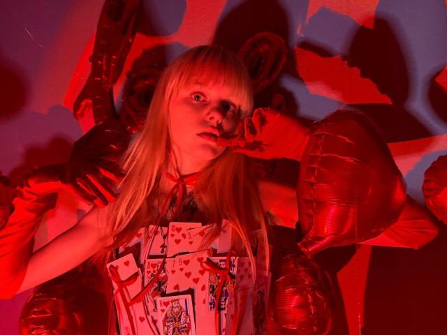 Lialit with blonde hair, wearing a red and white heart-patterned dress, has red-gloved hands near her face, under red lighting. Red bear-like balloons surround her.