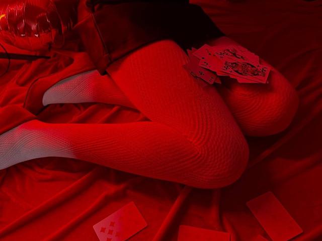 Lialits rote, strukturierte Fischgrätmuster-Beine sind auf einem roten Bett ausgebreitet, mit Spielkarten und einem rot verpackten Geschenk in ihrer Nähe. Ihre roten Absätze sind sichtbar..