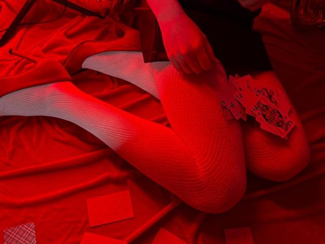 Lialit knielt, met rode fishnet leggings, en houdt speelkaarten vast, op een rode stof. Haar handen zijn in de buurt van haar schaamstreek, en er ligt een rood vierkant op het bed..