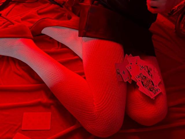 Las piernas de Lialit, cubiertas con medias de red rojas, están dobladas sobre una cama roja, mientras sostiene una mano de póker con reyes y reinas. Su top negro se ve ligeramente. Una carta roja está cerca..
