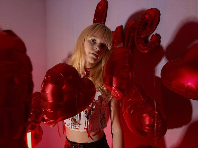 Lialit, met blond haar, draagt een witte crop top en houdt rode, hartvormige ballonnen vast. Ze heeft een slank postuur en een schattig gezicht, en staat in een kamer met roze verlichting..