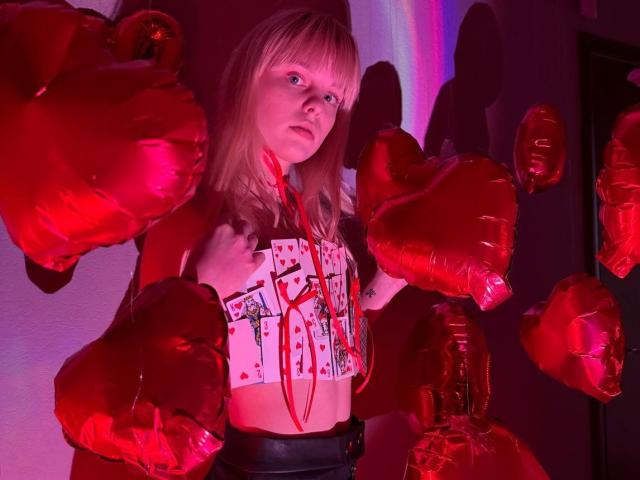 Lialit with blonde hair, wearing a card-themed top, surrounded by red heart balloons, has a serious expression, and is in a red-lit room, hands on her chest.