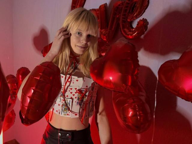Lialit, con cabello rubio, vestida con una camiseta corta con estampado de corazones, cintas rojas y pantalones negros, sosteniendo globos con forma de corazón rojos, sonriendo, en una habitación iluminada con luz roja..
