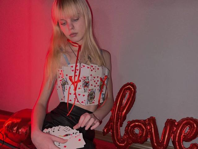 Lialit has blonde hair, wearing a heart-patterned top, holding cards, with a red string, and black skirt, next to red "love" balloons, in a red-lit room.