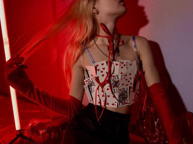 Lialit, con cabello rubio largo, vestida con un top azul de tirantes, sosteniendo una caja roja con estampado de corazones, guantes rojos y cintas rojas, con iluminación roja y azul. Se ven los senos..