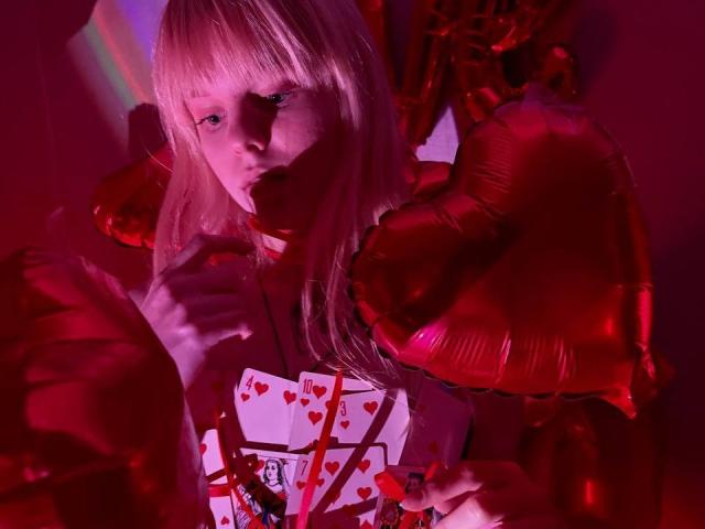 Lialit with blonde hair, red balloons, and a deck of heart cards, holding a red-wrapped gift, looking seductive with red lighting, showing a sexy, cute face.