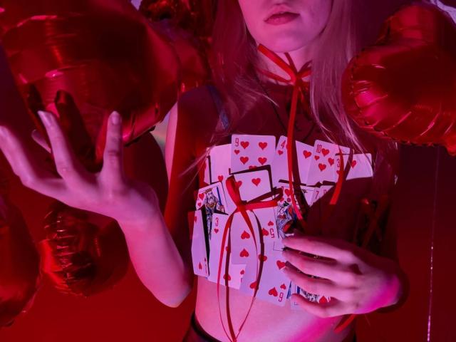 Lialit with red balloon fists, holding a deck of cards with hearts, wearing a red crop top and ribbon, under pink and red lighting. Boobs visible, face partially shown.