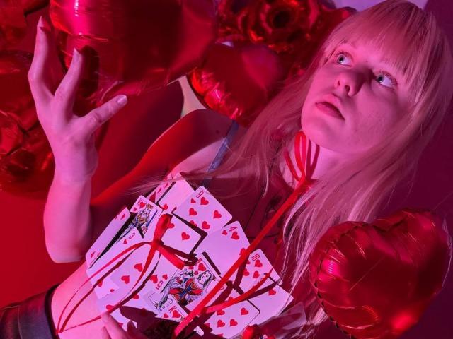 Lialit mit blonden Haaren, die rote Herzballons hält und eine Karte mit Assen und Herzsymbolen hat, trägt ein rotes Band um den Hals und blickt mit einem leicht überraschten Ausdruck nach oben..