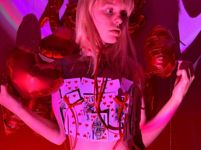Lialit, con el pelo rubio largo, vestida con una camiseta negra con un diseño, sosteniendo globos con forma de corazón rojos, tiene una expresión de puchero, bajo una iluminación rosa y roja, en una pose juguetona y sensual..