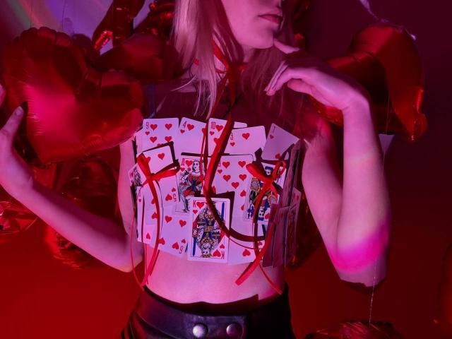 Lialit, con cabello rubio, sosteniendo globos en forma de corazón rojo, vistiendo una blusa cubierta de cartón con corazones rojos y la letra "X", cinturón negro, con un dedo en el labio, brazo rosa, iluminación roja..