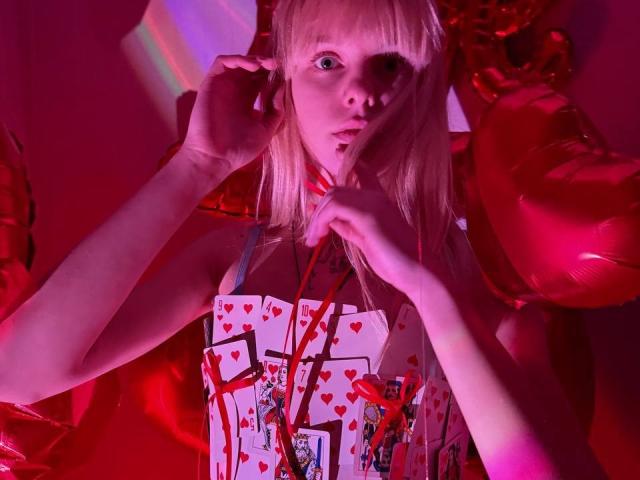 Lialit with blonde hair, red heart-patterned top, and red balloon, holding string, pink and red lighting, finger to mouth, looking curious, tits barely visible, cute, sexy.