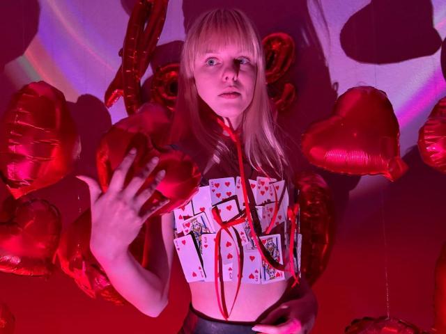 Lialit with blonde bangs, wearing a card-themed top, holding red heart balloons, has a serious expression, red lighting, and a ribbon in her hair, in a colorful, red-lit setting.