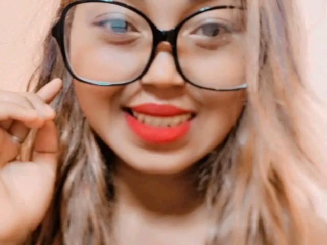 SousouHot with big black glasses, winking, and red lipstick, touching her blonde hair with a cute smile, showing her teeth. She's got a sexy, nerdy look.