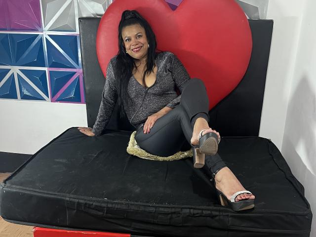 HellenaSexy, with black hair and red lipstick, sits on a black mattress, wearing a gray sparkly top, black pants, and silver heels, showing her tits and crossed legs.