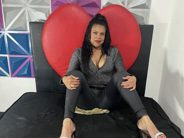 HellenaSexy, com cabelo preto preso em um rabo de cavalo, usando uma blusa cinza e calças cinzas justas, sentada em uma cama preta com um grande coração vermelho atrás dela, com as pernas abertas, mostrando sua vulva..